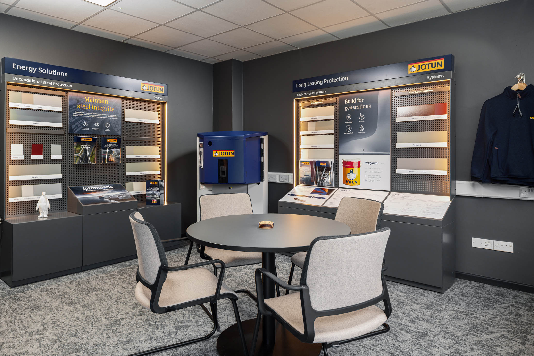 Modern showroom interior with Jotun product displays showcasing steel protection systems and coatings, featuring sample panels, branded literature, and a round meeting table with four upholstered chairs.